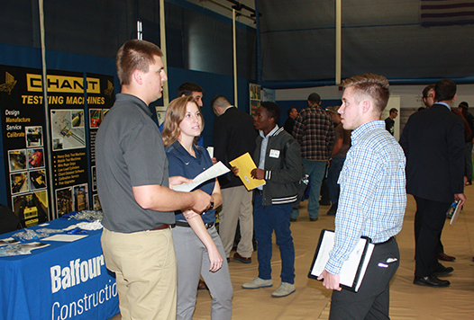 student holding notebook speaking with employer standing at a table at Career Fair in the gymnasium
