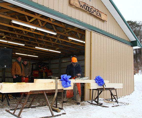 Alfred State President Dr. Skip Sullivan uses a chainsaw Wednesday to cut the wooden “ribbon” at a ceremony to celebrate the new Pioneer Woodsmen’s Club Barn.
