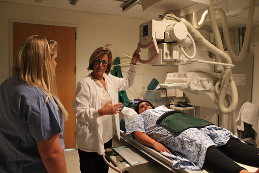 professor demonstrating medical equipment, female student watching and female student lying on medical equipment